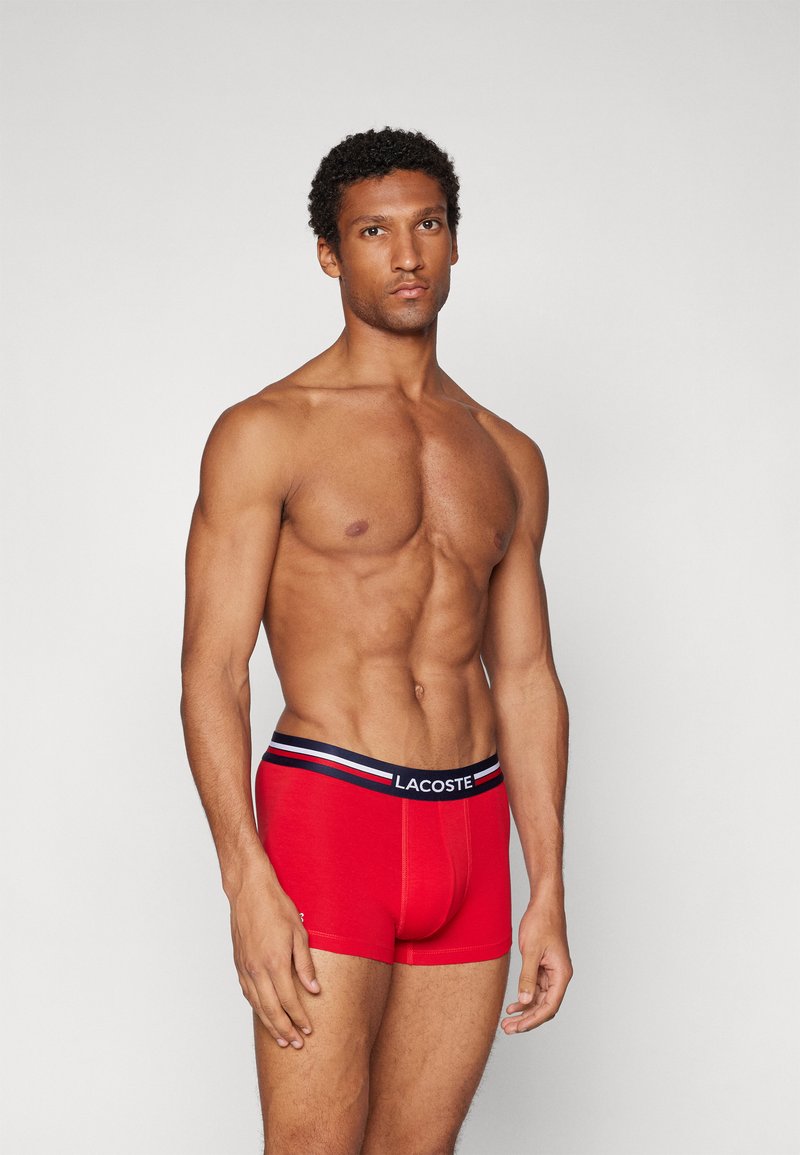 Boxer-culottes rouges en coton, dotés d'une taille noire avec des rayures blanches et rouges et du logo Lacoste. Texture lisse et design ajusté.