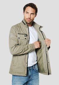 Khaki bunda se zipem vpředu, žebrovaným límcem a několika kapsami. Materiál je hladký s mírným leskem. Nošena přes bílou košili.