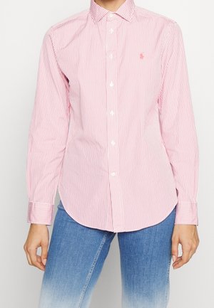 Person trägt ein rosa-weiß gestreiftes Hemd mit langen Ärmeln und Kragen sowie blaue Jeans.