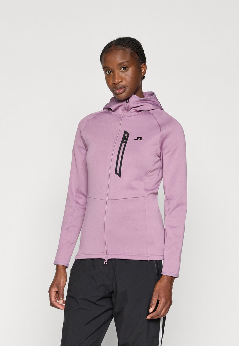 Sudadera con cremallera de color púrpura claro y diseño ajustado, con capucha y cremallera negra. Presenta un logotipo en el pecho y detalles de costura sin costuras en las mangas.