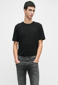 Νεαρός με κοντά σκούρα μαλλιά, φορώντας ένα απλό μαύρο μπλουζάκι και γκρι τζιν, στέκεται με τα χέρια στις τσέπες σε λευκό φόντο.