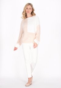 Lekki sweter dziewiarski o geometrycznym wzorze w kolorze białym i delikatnym brzoskwiniowym, z fakturowanymi wzorami i szerokimi rękawami, zestawiony z białymi spodniami.