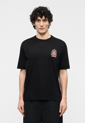 Junger Mann mit lockigem Haar trägt ein schwarzes T-Shirt mit kleinem rotem und grauem verziertem Wappen auf der linken Brust, steht vor schlichtem Hintergrund.