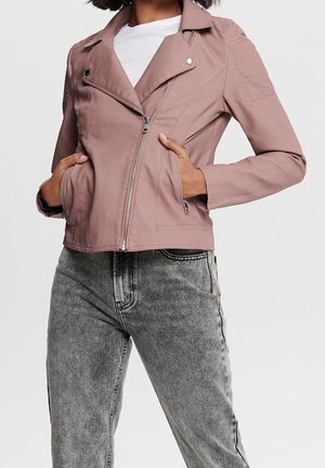 Femme portant une veste de motard mauve clair zippée avec un détail matelassé aux épaules et un jean gris délavé, mains dans les poches de la veste.