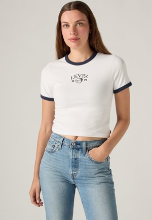 Jeune femme portant un t-shirt court blanc Levi's avec un liseré bleu marine et un jean taille haute bleu clair, debout avec une main dans la poche.