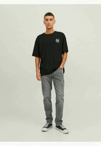 Zwart t-shirt met korte mouwen met een logo op de linkerborst, gecombineerd met grijze jeans en zwarte sneakers, staand tegen een neutrale achtergrond.
