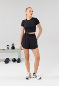 Zwarte cropped sporttop met bijpassende korte broek met elastische tailleband. Zilveren en zwarte hardloopschoenen maken de outfit compleet. Fitnessapparatuur op de achtergrond.
