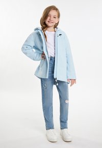 Ljusblå zip-up jacka med huva och broderad detalj, tillsammans med slitna blå jeans med en fjärilsmotiv och vita sneakers.