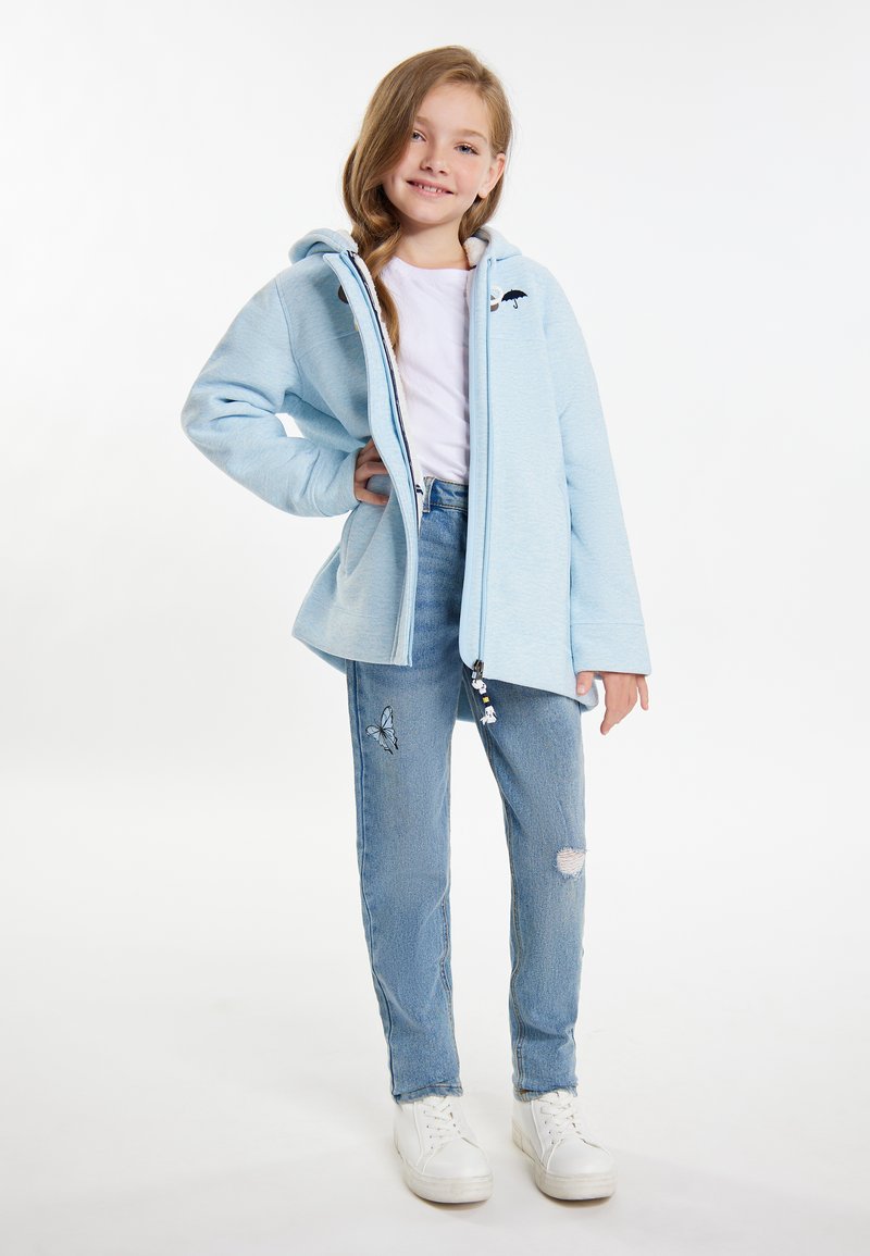 Ljusblå zip-up jacka med huva och broderad detalj, tillsammans med slitna blå jeans med en fjärilsmotiv och vita sneakers.