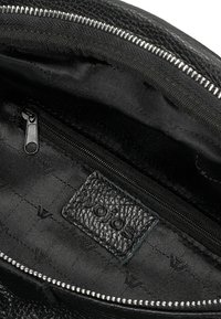 Vnitřek černé kožené tašky ukazuje zipovou kapsu, texturovanou látku a značkovou etiketu s jemným leskem. Detail stříbrného zipu.