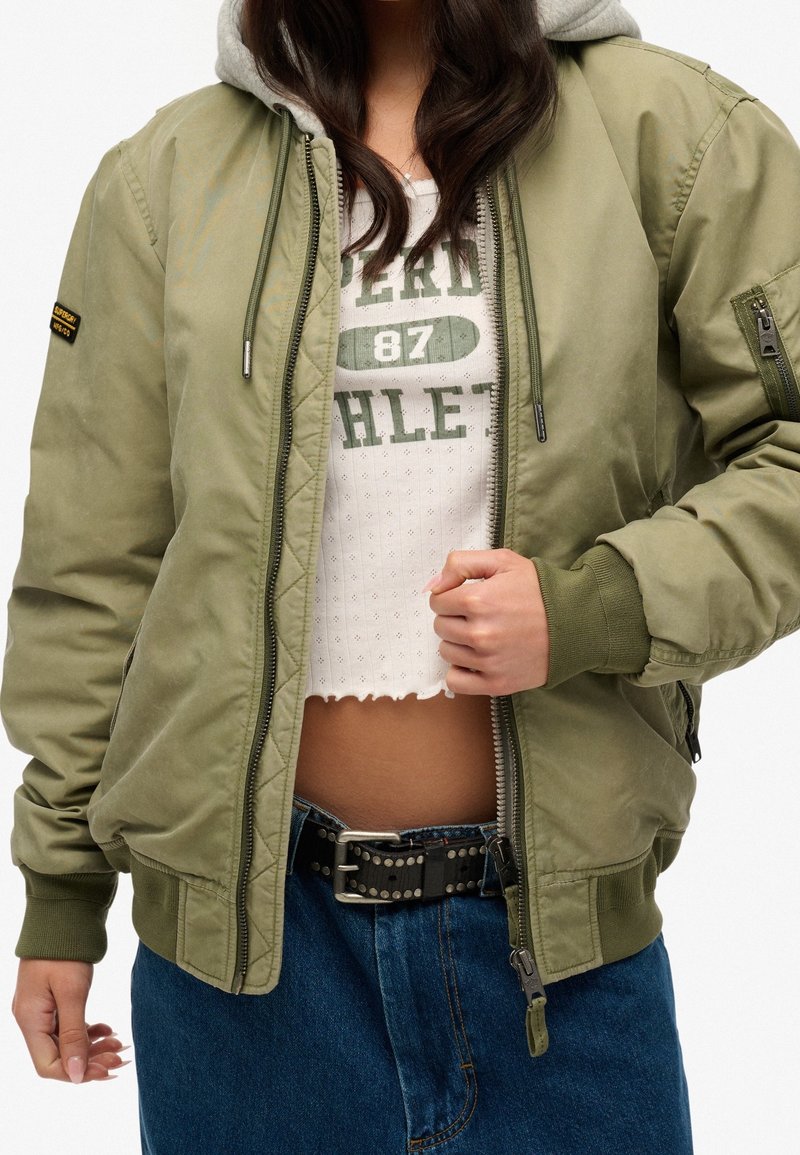 Groene bomberjack met een grijze hoodie, voorzien van ritszakken, ribgebreide manchetten en een rits aan de voorkant. Draagt over een witte crop top en een spijkerrok.