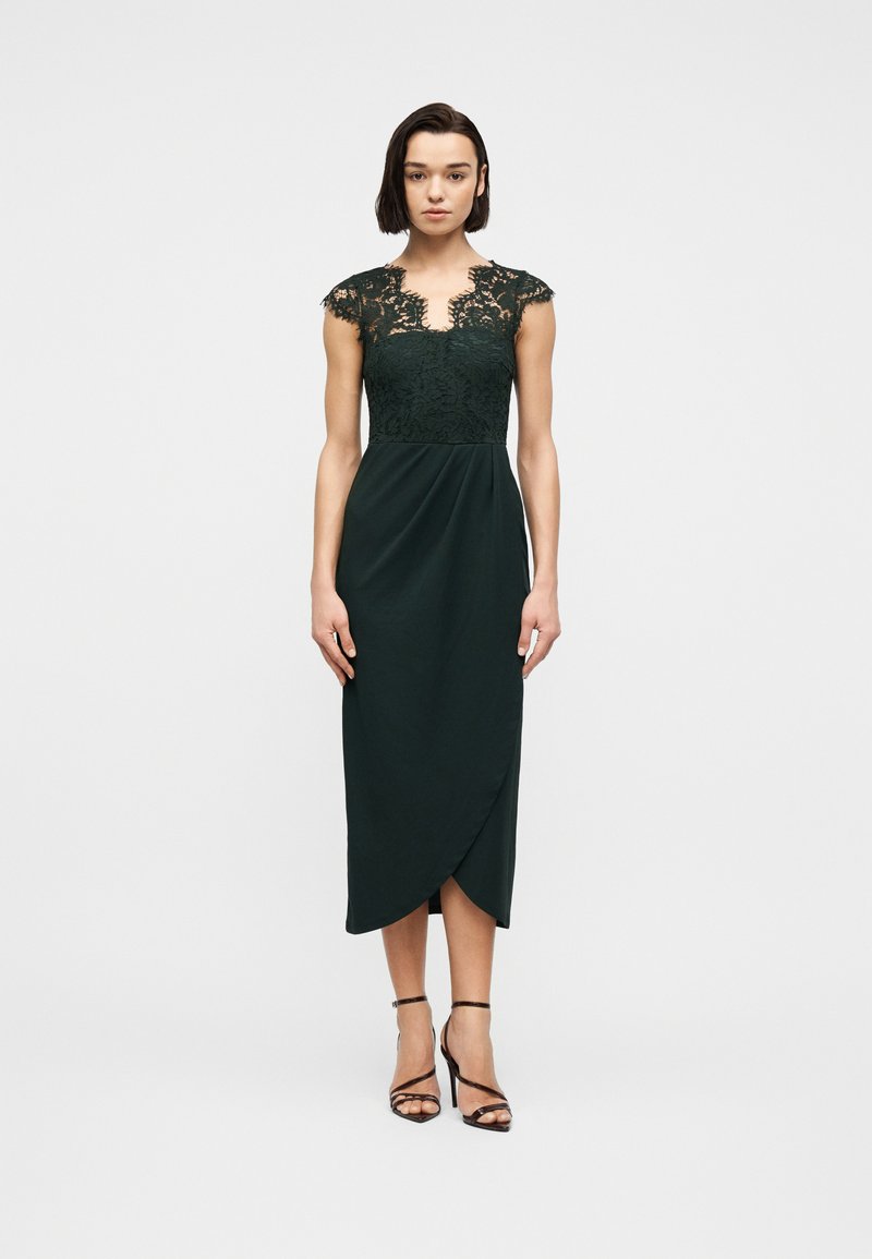 Γυναίκα που στέκεται, φοράει εφαρμοστό midi φόρεμα σε σκούρο πράσινο με δαντελωτές μανσέτες-καπέλα στα μανίκια και μαύρα ψηλοτάκουνα πέδιλα με λουράκια, σε λευκό φόντο.