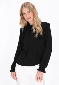 Schwarze Bluse mit langen Ärmeln, Rüschenkragen und -manschetten, in lockerer Passform und aus leichtem Stoff. Das Model trägt weiße Hosen.