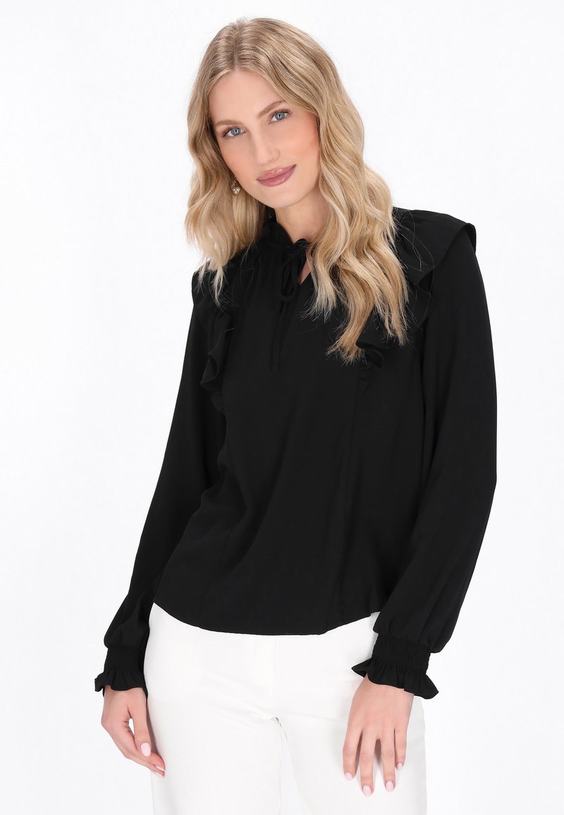 Schwarze Bluse mit langen Ärmeln, Rüschenkragen und -manschetten, in lockerer Passform und aus leichtem Stoff. Das Model trägt weiße Hosen.
