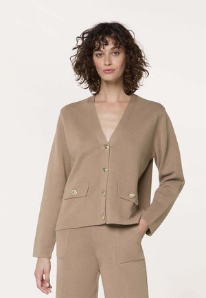 Vrouw met krullend haar, gekleed in een beige gebreid vest met knopen en voorzakken, en een bijpassende broek, poserend met één hand in de zak.