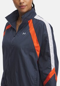 Navy blue zip-up jacket with orange and white stripes. Features a high collar and lightweight, textured fabric. Under Armour logo displayed.