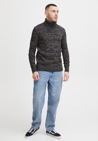 Mørk teksturert polokrage genser med ribbestrikket kant, iført lyseblå jeans og svarte joggesko. Genseren har en liten logo detalj.