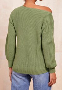 Pull en maille vert olive, avec un design épaules dénudées, des poignets côtelés et un motif texturé. Associé à un jean bleu clair.