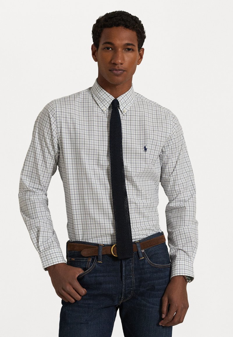 Camisa blanca de botones con un patrón de cuadros en azul y verde, con un pequeño logo, combinada con jeans oscuros y una corbata texturizada color marino.
