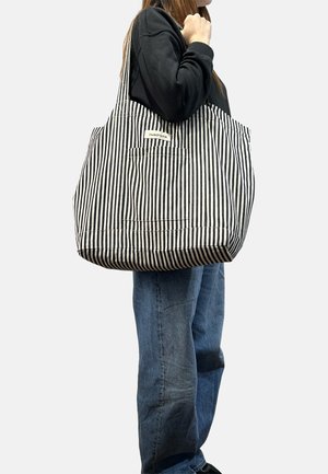 Person wearing black sweatshirt and blue jeans carries large black-and-white vertical striped tote bag on shoulder.