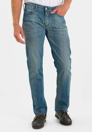 Man die vervaagde blauwe jeans en bruine veterschoenen draagt, staande met één hand in zijn zak tegen een effen achtergrond.