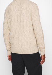 Suéter de punto beige con un patrón de diamante texturizado, cuello redondo y puños acanalados; combinado con pantalones oscuros.