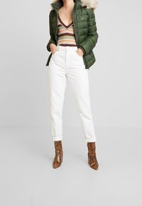 Kvinna iklädd en grön puffjacka med pälshem, randig V-ringad topp, vita högmidjade byxor och bruna ormskinnsankelskängor.