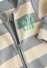 Grå og creme-striped lynlåse onesie med ribbet tekstur. Har forhøjede grønne "MILK CLUB" bogstaver og en grå lynlås.