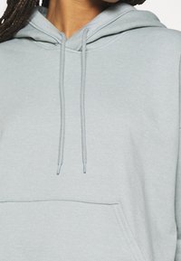 Jasnoszara bluza z kapturem, z kieszenią z przodu, sznurkami i miękką fakturą. Posiada klasyczny krój oraz ściągacze przy rękawach.