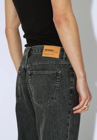 Osoba w wyblakłych czarnych dżinsach z denimu z skórzaną naszywką marki DRDENIM na pasku, w połączeniu z czarnym topem bez rękawów.