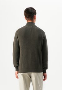 Pullover di maglia verde scuro con colletto alto, texture a coste e maniche lunghe. Abbinato a pantaloni di colore chiaro.