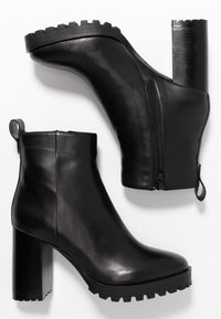 Mustat nahkaiset nilkkasukat, joissa on paksut, sahalaitapohjat, korkea tukikorko ja sivuvetoketjut; takana on vetotuli.