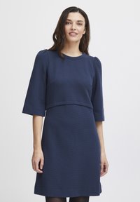 Mörkblå klänning i texturerad tyg, korta ärmar och en strukturerad bystdesign. Sömmar betonar midjan och ärmarna.
