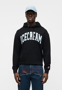 Μαύρο hoodie με την επιγραφή "ICECREAM" σε μπλε γράμματα, συνδυασμένο με μπλε τζιν παντελόνι που διαθέτει κόκκινο και άσπρο διακοσμητικό σχέδιο γάτας και λεπτομέρεια στην τσέπη.