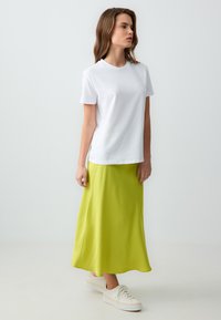 Weißes T-Shirt mit kurzen Ärmeln kombiniert mit einem langen, lebhaft gelben Satinrock und weißen Plateauschuhen. Sanfte Texturen und lockere Passform.