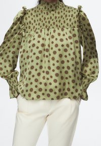 Blusa verde con motivo floreale in rosso e verde scuro. Presenta un collo alto smockato e maniche lunghe puff con accenti arricciati.