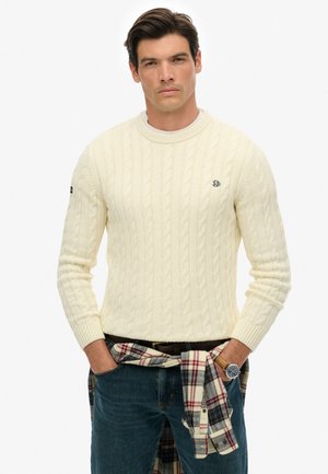 Jersey de punto trenzado color crema con escote redondo, llevado sobre una camisa blanca. Combinado con jeans de mezclilla y una camisa de cuadros atada a la cintura.