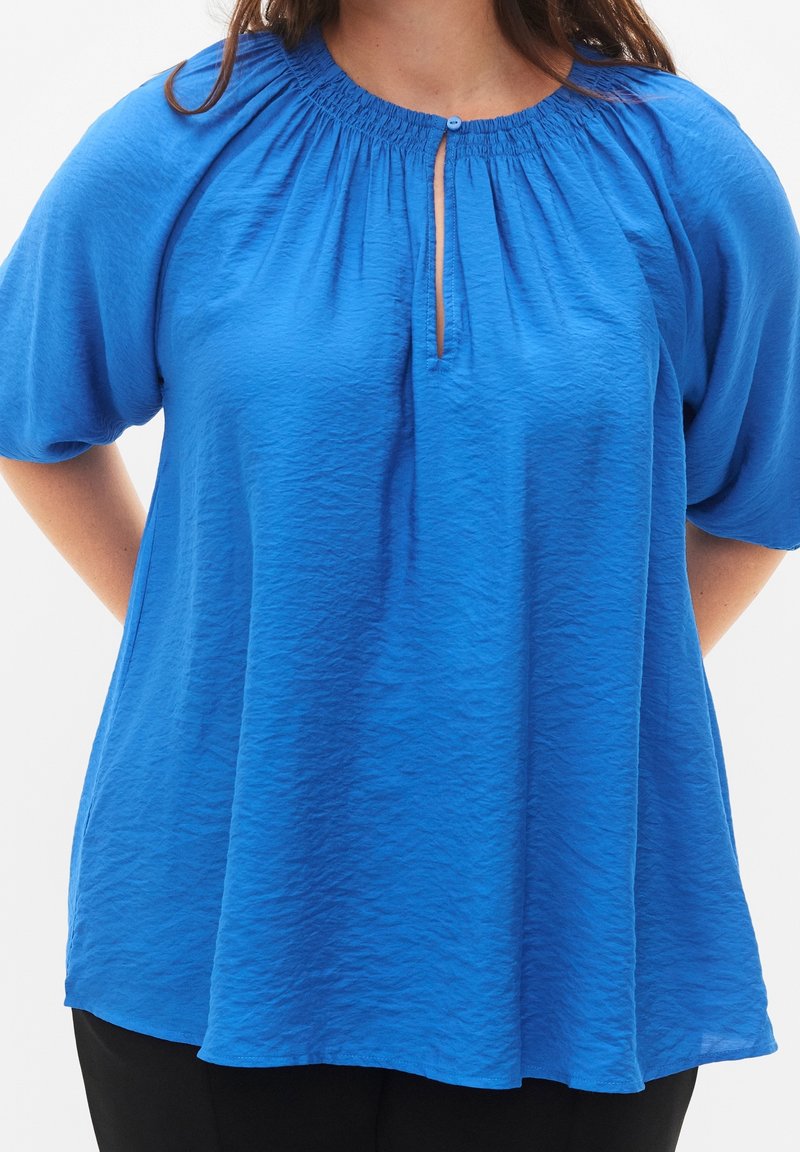 Blusa azul de manga corta hecha de un tejido liviano y arrugado, con un escote fruncido y un cierre de botón centrado en la abertura.