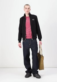 Casaco preto com fecho, por cima de uma t-shirt rosa, combinado com jeans azuis largos e ténis cinzentos. Segura uma bolsa de mão bege. Fundo branco liso.