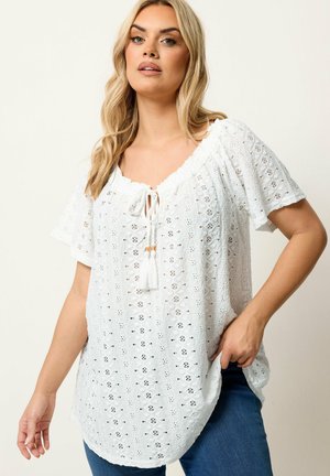 Kobieta o blond włosach, ubrana w białą bluzkę z krótkim rękawem z ażurowym wzorem i wiązaniem z przodu oraz niebieskie dżinsy, pozująca na neutralnym tle.