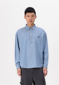 Cămașă din denim albastru deschis cu mânecă lungă, având guler ascuțit, plachetă cu nasturi și un buzunar pe piept cu un logo. Textură netedă, croială relaxată.