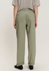 Persoon in lichtgroene omgeslagen broek, beige T-shirt met korte mouwen en zwarte platte schoenen, gezien van achteren op een effen achtergrond.