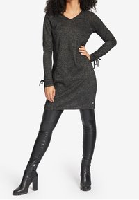 Schwarzes gestricktes Kleid mit langen Ärmeln und V-Ausschnitt. Verfügt über Kordelzüge an den Manschetten und ein Logo-Detail nahe dem Saum. Kombiniert mit schwarzen Lederleggings und Stiefeln.