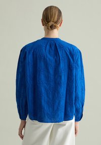 Blusa blu ricamata con maniche lunghe, caratterizzata da intricate fantasie floreali e un colletto rotondo. Tessuto testurizzato con una vestibilità ampia.
