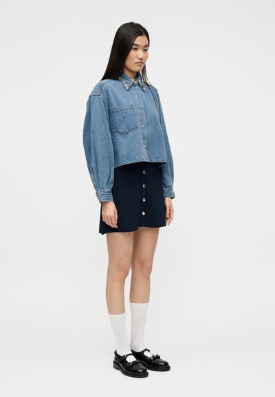 Νεαρή γυναίκα με υπερ μεγέθη τζιν πουκάμισο με διακοσμημένο γιακά, σκούρα μπλε φούστα με κουμπιά, λευκές κάλτσες και μαύρα παπούτσια Mary Jane, που στέκεται πλευρικά.