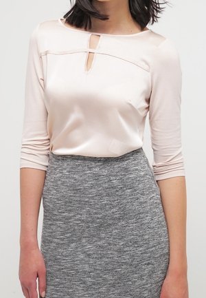 Kobieta w bladoróżowej satynowej bluzce z wycięciem na dekolcie i rękawami do połowy łokcia, w połączeniu z wysokim stanem szarej dzianinowej spódnicy.