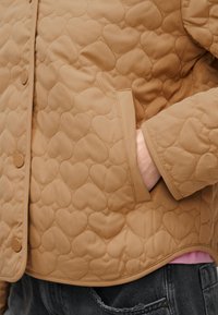 Ανοιχτό καφέ μπουφάν με καπιτονέ σχεδίαση σε σχήμα καρδιάς, πλαϊνή τσέπη και κουμπιά με τρουκς, φορεμένο πάνω από μαύρο τζιν.