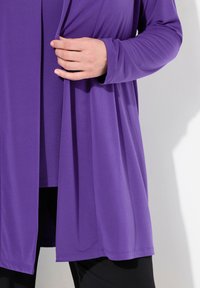 Lila langärmlige Strickjacke aus leichtem Stoff, mit offenem Vorderteil, glatter Textur, über einem dunkleren Oberteil und schwarzen Hosen getragen.