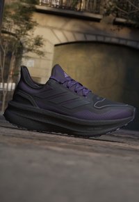 Sort og lilla Adidas løbesko med boost-sål, placeret udendørs på en brostensbelægning nær en stensbro.