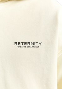 Cremfarbener Sweatshirt aus weichem Material mit schwarzem Text auf dem Rücken, der "RETERNITY CREATIVE DEPARTMENT" liest.